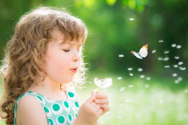 Young girl blowing dandelion seeds as a butterfly flutters nearby, symbolizing wonder and connection – Little Guru & Co. FAQ