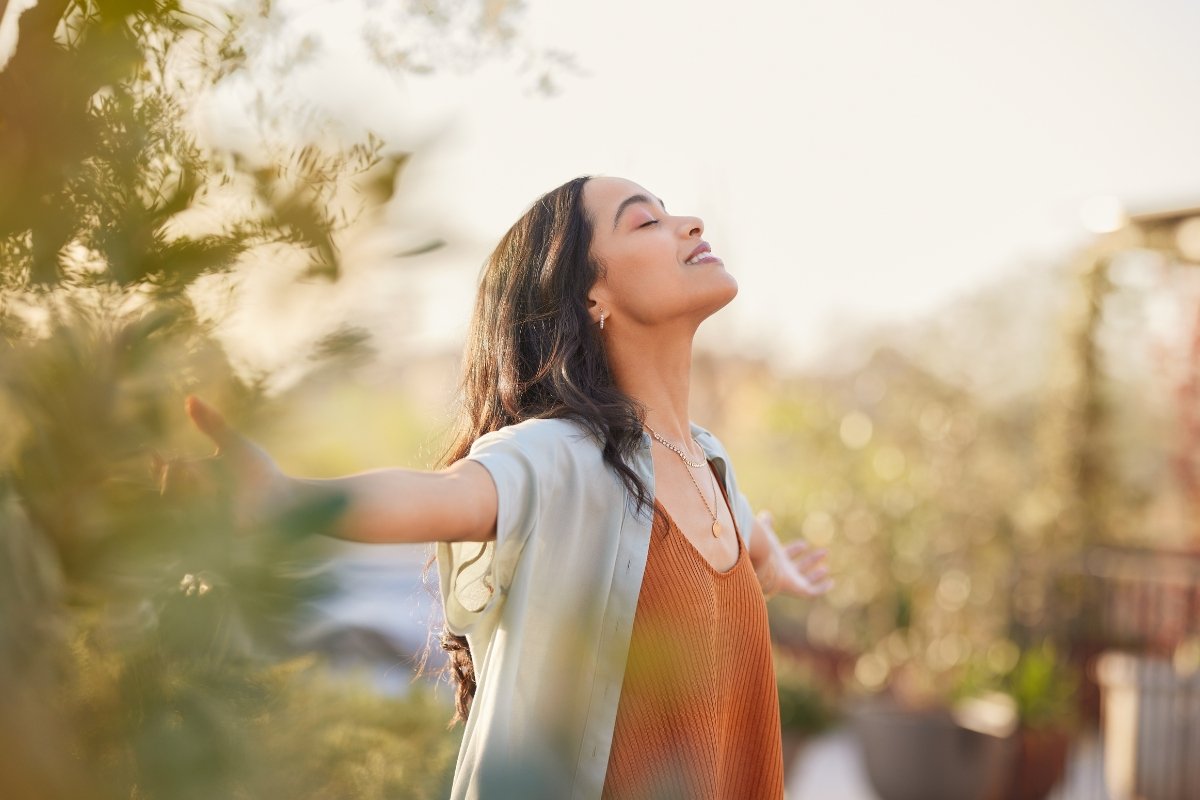 Woman standing outdoors with eyes closed and arms open, breathing deeply in sunlight, reflecting becoming present as a parent through mindful awareness and calm presence.