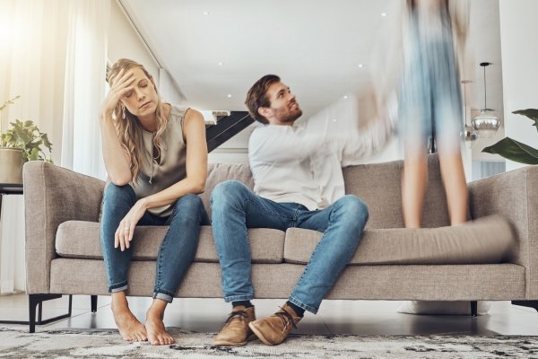 Two parents sitting on a couch looking tired and overwhelmed while a child jumps nearby, illustrating the challenges of staying present during everyday parenting moments.