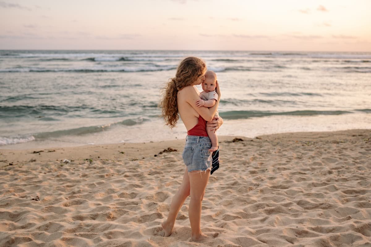 Re-membering: Sacred Wholeness as a Parenting Practice Adult holding a baby at the shoreline, facing the ocean in a moment of grounding and continuity.