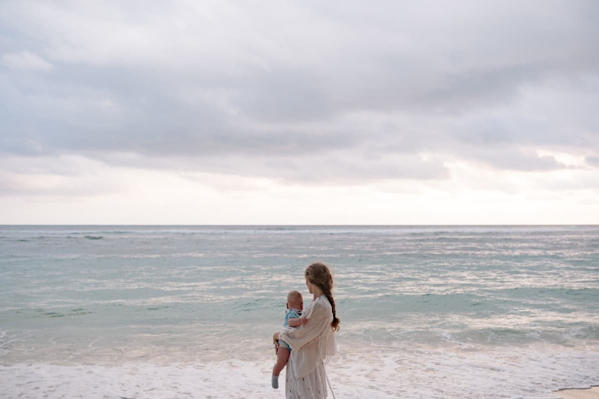 Re-membering: Sacred Wholeness as a Parenting Practice Adult holding a baby while facing the ocean, symbolizing return, memory, and the wider world.