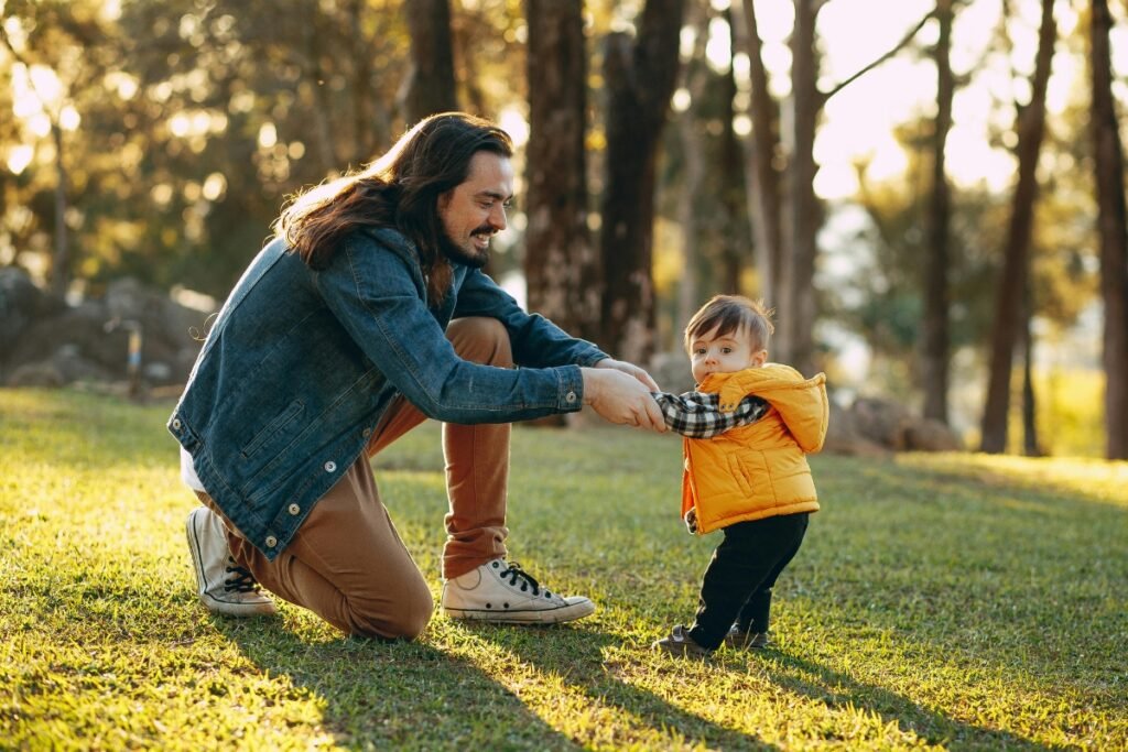 Father kneeling in a sunlit park holding his toddler’s hands as the child takes early steps, symbolizing supportive and soulful parenting